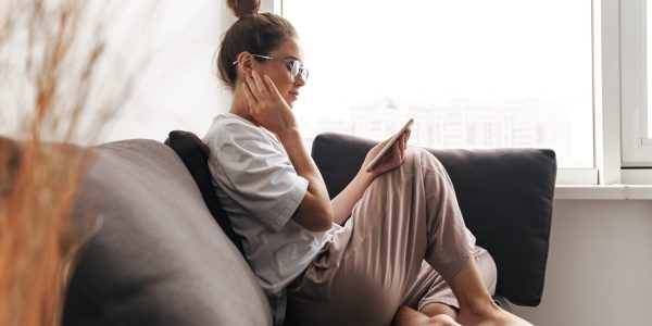 Image of concentrated nice woman in eyeglasses using cellphone while sitting on sofa at living room